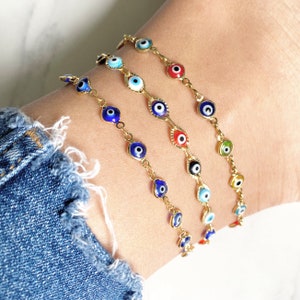 May include: Three gold-tone chain bracelets featuring small, circular evil eye charms in various colors including blue, red, white, green, yellow, and black. The bracelets are worn together, showcasing a delicate link design with multiple eye motifs.