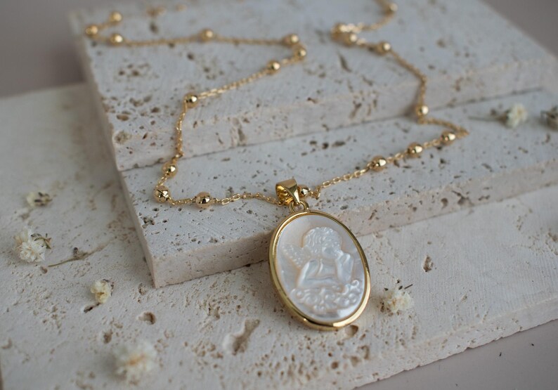 Puede incluir: Un collar de tono dorado con una cadena delicada de peque&ntilde;as cuentas esf&eacute;ricas y un colgante ovalado. El colgante contiene un camafeo blanco iridiscente tallado con la figura de un querub&iacute;n, engastado en un marco de metal dorado pulido.