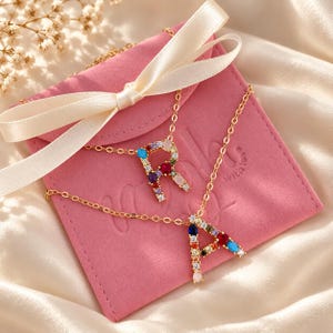 May include: Two gold-tone necklaces featuring letter pendants R and A, each embellished with multi-colored gemstones including red, blue, and clear stones. The necklaces are displayed on a pink velvet pouch with a cream ribbon bow.
