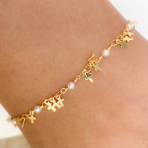 Puede incluir: Una pulsera de cadena en tono dorado que presenta pequeños dijes de cruz colgantes y cuentas esféricas blancas intercaladas. Los delicados eslabones metálicos están conectados con detalles de alambre enrollado, creando un patrón continuo de cruces doradas y cuentas blancas.