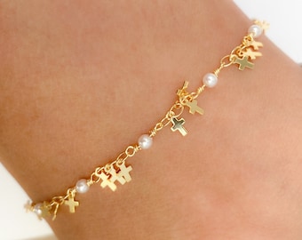 May include: A gold-toned chain bracelet featuring small, dangling cross charms and interspersed white spherical beads. The delicate metal links are connected with wire-wrapped details, creating a continuous pattern of alternating gold crosses and white beads along the length of the jewelry piece.