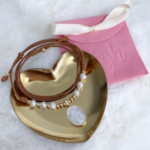 K&ouml;nnte beinhalten: Eine Halskette aus braunem Lederband mit einer Reihe wei&szlig;er S&uuml;&szlig;wasserperlen und kleinen goldfarbenen Perlen. Die Kette enth&auml;lt einen ovalen Anh&auml;nger mit einer wei&szlig;en Perlmutt-Einlage, die eine religi&ouml;se Figur darstellt, auf einem goldenen Herz.