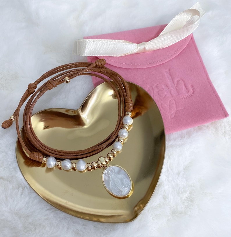 K&ouml;nnte beinhalten: Eine Halskette aus braunem Lederband mit einer Reihe wei&szlig;er S&uuml;&szlig;wasserperlen und kleinen goldfarbenen Perlen. Die Kette enth&auml;lt einen ovalen Anh&auml;nger mit einer wei&szlig;en Perlmutt-Einlage, die eine religi&ouml;se Figur darstellt, auf einem goldenen Herz.