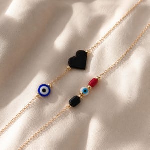 Puede incluir: Dos cadenas delicadas en tono dorado con dijes decorativos. Una cadena muestra un coraz&oacute;n negro y una cuenta de ojo azul. La segunda cadena presenta una cuenta cil&iacute;ndrica roja, una peque&ntilde;a cuenta de ojo azul y una cuenta rectangular negra.