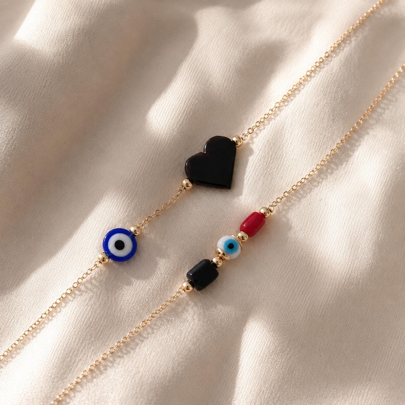 Puede incluir: Dos cadenas delicadas en tono dorado con dijes decorativos. Una cadena muestra un coraz&oacute;n negro y una cuenta de ojo azul. La segunda cadena presenta una cuenta cil&iacute;ndrica roja, una peque&ntilde;a cuenta de ojo azul y una cuenta rectangular negra.
