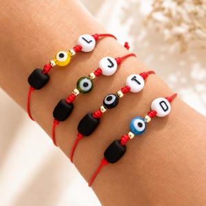 Puede incluir: Cuatro pulseras de hilo rojo con cuentas rectangulares negras, pequeños espaciadores dorados y cuentas redondas con diseños de ojo turco en amarillo, verde, negro y azul. Cada pulsera incluye una cuenta blanca con una letra negra L, J, T o D.