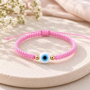 K&ouml;nnte beinhalten: Ein Armband aus rosa geflochtener Kordel mit einer zentralen, runden Glasperle in Form eines Schutzauges in Wei&szlig;, Hellblau und Schwarz, flankiert von zwei kleinen, goldfarbenen Metallperlen. Das Armband hat einen verstellbaren Schiebeknoten.