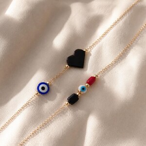 Puede incluir: Dos cadenas delicadas en tono dorado con dijes decorativos. Una cadena muestra un coraz&oacute;n negro y una cuenta de ojo azul. La segunda cadena presenta una cuenta cil&iacute;ndrica roja, una peque&ntilde;a cuenta de ojo azul y una cuenta rectangular negra.