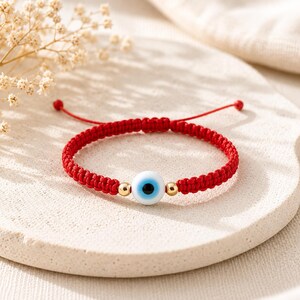 K&ouml;nnte beinhalten: Ein Armband aus roter geflochtener Kordel mit einer zentralen, runden, wei&szlig;en Perle in Form eines b&ouml;sen Blicks mit blauer und schwarzer Iris. Das Armband ist mit zwei kleinen goldfarbenen Metallkugeln an den Seiten der Perle und verstellbaren Enden versehen.