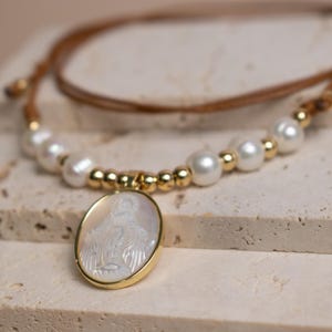K&ouml;nnte beinhalten: Eine Halskette mit braunem Lederband, wei&szlig;en Perlen und kleinen goldfarbenen K&uuml;gelchen. Das Herzst&uuml;ck ist ein ovaler Anh&auml;nger mit einer wei&szlig;en, schimmernden Perlmutt-Einlage, die eine Figur darstellt und in einen goldfarbenen Metallrahmen gefasst ist.