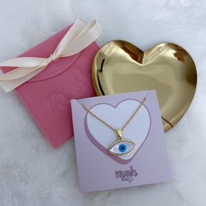 May include: A gold-tone chain necklace with an eye-shaped pendant featuring a white mother-of-pearl inlay and a central blue pupil. The necklace is displayed on a square card with a heart graphic and the text "nyah", alongside a pink pouch.