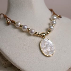 K&ouml;nnte beinhalten: Eine Halskette mit einem zentralen ovalen Anh&auml;nger, der eine geschnitzte religi&ouml;se Figur aus Perlmutt in einem goldfarbenen Rahmen zeigt. Die Kette besteht aus wei&szlig;en S&uuml;&szlig;wasserperlen und kleinen goldfarbenen Perlen an einer braunen Kordel.