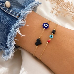 Puede incluir: Dos pulseras de cadena en tono dorado. Una presenta una cuenta de ojo turco azul oscuro y una cuenta negra en forma de coraz&oacute;n. La segunda pulsera incluye una peque&ntilde;a cuenta rectangular negra, una cuenta de ojo turco azul claro y una cuenta esf&eacute;rica roja.