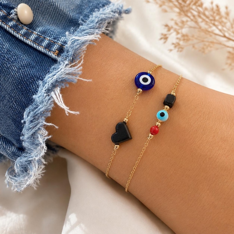 Puede incluir: Dos pulseras de cadena en tono dorado. Una presenta una cuenta de ojo turco azul oscuro y una cuenta negra en forma de coraz&oacute;n. La segunda pulsera incluye una peque&ntilde;a cuenta rectangular negra, una cuenta de ojo turco azul claro y una cuenta esf&eacute;rica roja.