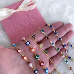 Puede incluir: Un conjunto de cinco pulseras de cadena dorada con dijes de ojo turco de colores. Cada pulsera presenta una combinaci&oacute;n de colores diferente de azul, verde, rojo, naranja y rosa. Las pulseras se muestran en una mano sobre un fondo blanco.