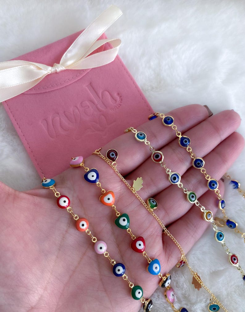 Puede incluir: Un conjunto de cinco pulseras de cadena dorada con dijes de ojo turco de colores. Cada pulsera presenta una combinaci&oacute;n de colores diferente de azul, verde, rojo, naranja y rosa. Las pulseras se muestran en una mano sobre un fondo blanco.