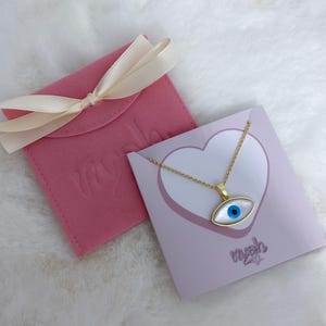 May include: A gold-tone chain necklace with an eye-shaped pendant featuring a white mother-of-pearl inlay and a blue center pupil. The necklace is displayed on a light purple card with a heart graphic and the text nyah, next to a pink pouch.