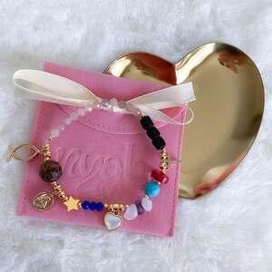 May include: A gold charm bracelet with a variety of colorful beads, including pink, red, blue, turquoise, and purple. The bracelet also features a gold star charm, a gold heart charm, a gold fish charm, and a gold religious charm. The bracelet is displayed on a pink velvet pouch with the word "Myah" written on it.