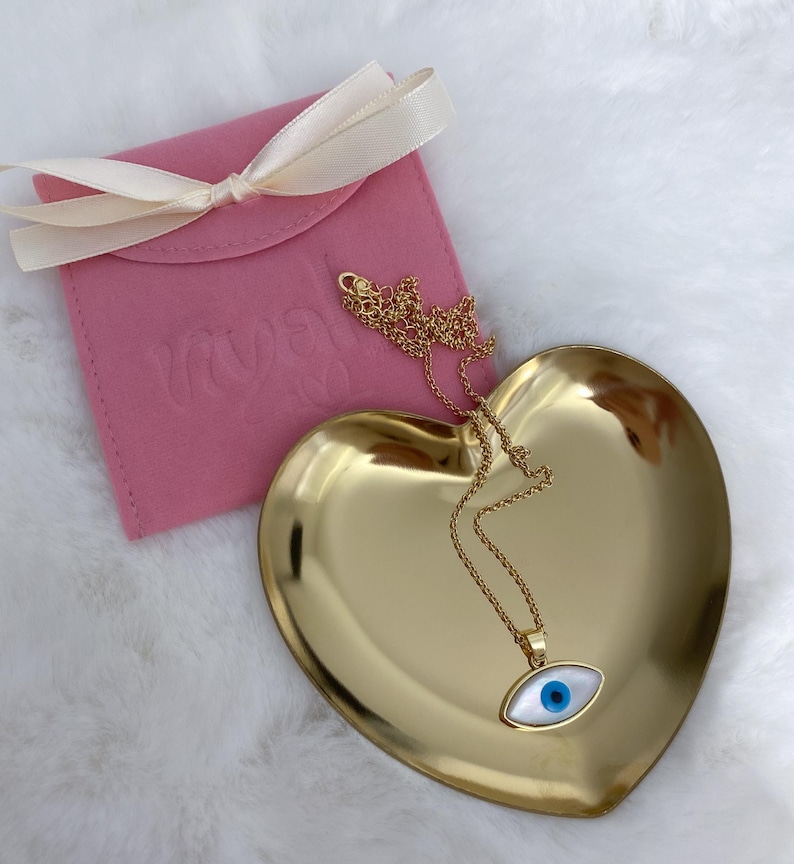 May include: A gold-tone chain necklace with an evil eye pendant featuring a white oval base and a central blue pupil. The necklace rests on a gold-tone heart-shaped dish next to a pink fabric pouch tied with a cream-colored ribbon.