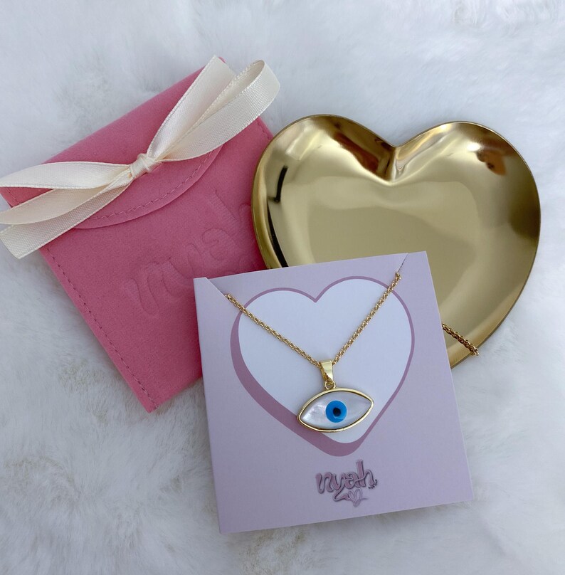 May include: A gold chain necklace with an eye-shaped pendant featuring a white mother-of-pearl inlay and a central blue pupil. The necklace is displayed on a light purple card with the text 'nyah' next to a gold heart-shaped dish and a pink pouch.