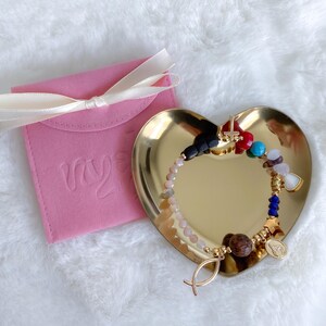 May include: A gold heart-shaped dish with a colorful beaded bracelet featuring a cross, fish, and star charms. The bracelet is on a pink velvet pouch with the word "my" embossed on it.