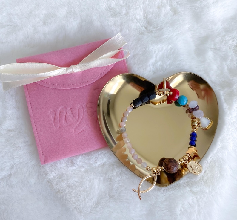 May include: A gold heart-shaped dish with a colorful beaded bracelet featuring a cross, fish, and star charms. The bracelet is on a pink velvet pouch with the word "my" embossed on it.