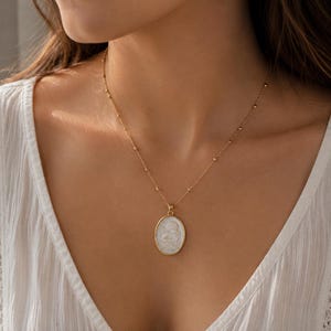Puede incluir: Un collar de cadena dorada con un delicado dise&ntilde;o de cuentas sat&eacute;lite y un colgante ovalado. El colgante consiste en una incrustaci&oacute;n de n&aacute;car blanco iridiscente engastada en un marco de metal dorado pulido, con un relieve tallado.