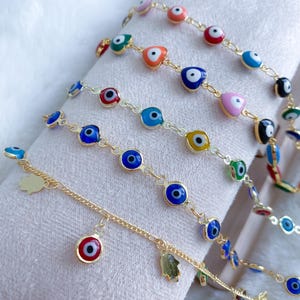 Puede incluir: Un conjunto de seis pulseras de cadena dorada con dijes de ojo azul maligno. Las pulseras presentan acentos de diferentes colores, incluyendo rosa, rojo, amarillo y verde. Una pulsera tiene un dije con una mano y un dije con un ojo maligno.