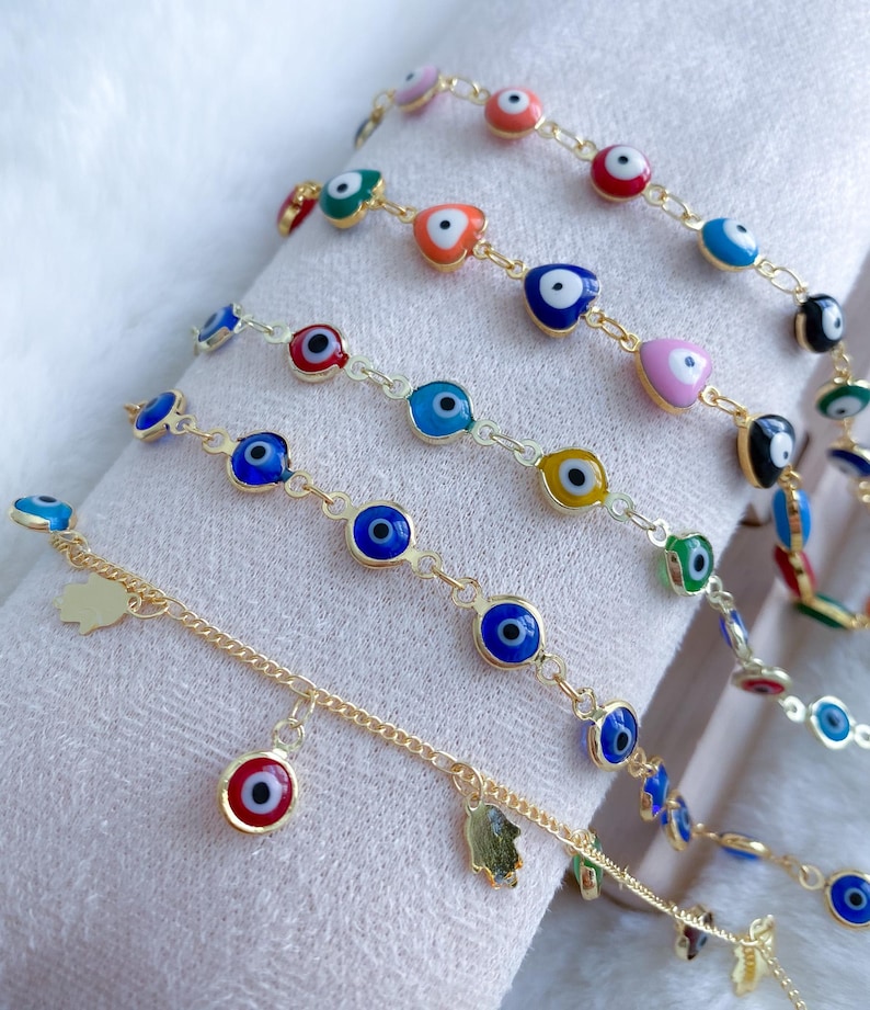 Puede incluir: Un conjunto de seis pulseras de cadena dorada con dijes de ojo azul maligno. Las pulseras presentan acentos de diferentes colores, incluyendo rosa, rojo, amarillo y verde. Una pulsera tiene un dije con una mano y un dije con un ojo maligno.
