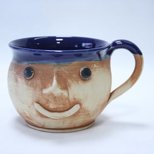 Könnte beinhalten: Eine handgefertigte Keramik-Tasse mit einem lächelnden Gesichtsdesign. Die Tasse hat einen blauen Innenraum und Rand, mit einem beigen und braunen Gesicht. Die Augen sind dunkelblau und der Griff ist ebenfalls blau. Eine einzigartige und skurrile Ergänzung für jede Küche.