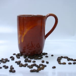 Könnte beinhalten: Eine braune Keramiktasse mit gelber Glasur steht auf einer weißen Oberfläche, umgeben von Kaffeebohnen.