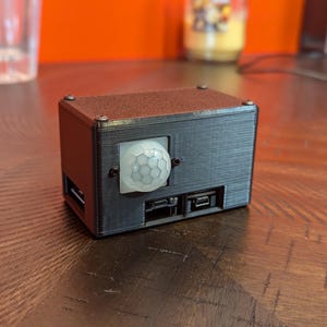 May include: A black, rectangular box with a white, round motion sensor on the front. The box has a textured surface and is sitting on a wooden table.