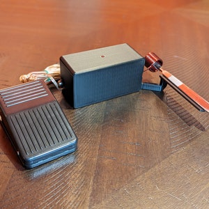 May include: A black foot pedal switch with a black box and a red light. The box has a copper coil and a metal rod attached to it.