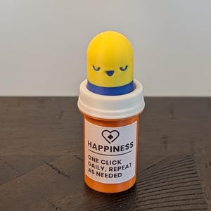 May include: A novelty pill bottle with a yellow, smiling capsule-shaped top. The orange bottle has a white label that reads "HAPPINESS" and "ONE CLICK DAILY, REPEAT AS NEEDED."