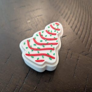 May include: A white, Christmas tree-shaped cookie cutter with red icing stripes and green sprinkles. The cookie cutter has a white border and is set against a dark wood grain surface.