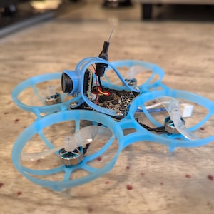 May include: A blue and black drone with four propellers and a camera. The drone is sitting on a light brown surface.