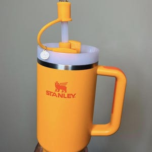 May include: A bright orange Stanley Flowstate H2.0 mug with a handle and a clear, gem-shaped topper. The mug has a white rim and a silver band. The Stanley logo is printed in orange. The mug is designed for cold beverages.