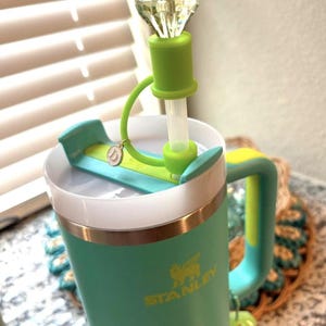 May include: A teal Stanley tumbler with a green handle and accents. The lid features a green straw and a decorative, clear, diamond-shaped topper. A small charm hangs from the lid. The Stanley logo is visible in gold.