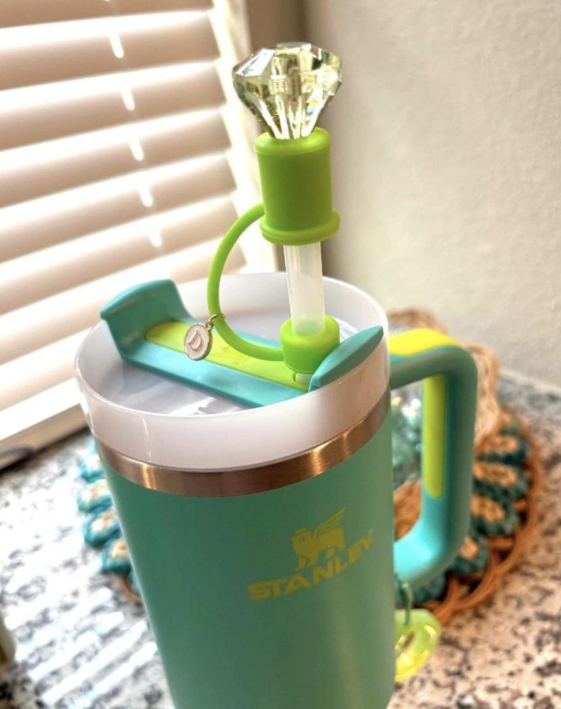 May include: A teal Stanley tumbler with a green handle and accents. The lid features a green straw and a decorative, clear, diamond-shaped topper. A small charm hangs from the lid. The Stanley logo is visible in gold.