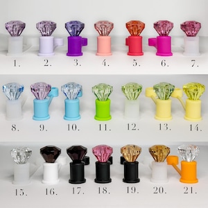 May include: A collection of twenty-one decorative bottle stoppers, each featuring a faceted gem-like top in various colors. The stoppers have a cylindrical base and a handle. Colors include clear, purple, pink, red, and yellow.