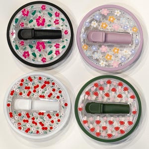 Pode incluir: Quatro recipientes redondos e transparentes com tampas decorativas. Cada tampa apresenta um design diferente: flores de hibisco rosa, flores coloridas, cerejas vermelhas e cogumelos vermelhos. Cada recipiente cont&eacute;m um objeto retangular colorido.