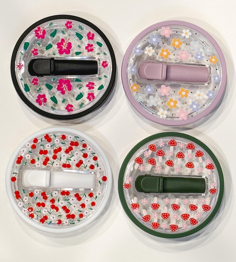Pode incluir: Quatro recipientes redondos e transparentes com tampas decorativas. Cada tampa apresenta um design diferente: flores de hibisco rosa, flores coloridas, cerejas vermelhas e cogumelos vermelhos. Cada recipiente cont&eacute;m um objeto retangular colorido.