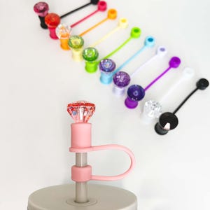May include: A collection of colorful bottle stoppers with jewel-like tops and attached silicone loops. The stoppers are in various colors, including pink, red, orange, yellow, green, blue, purple, white, and black. The stoppers are displayed on a white surface.