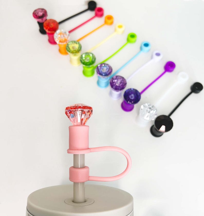 May include: A collection of colorful bottle stoppers with jewel-like tops and attached silicone loops. The stoppers are in various colors, including pink, red, orange, yellow, green, blue, purple, white, and black. The stoppers are displayed on a white surface.