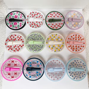 Pode incluir: Uma cole&ccedil;&atilde;o de doze rolhas de garrafa redondas com v&aacute;rios desenhos decorativos. Cada rolha tem uma cor e um padr&atilde;o diferentes, incluindo flores, cerejas, cogumelos e morangos. As rolhas s&atilde;o feitas de pl&aacute;stico transparente e t&ecirc;m uma pega.