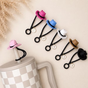 May include: A collection of straw hat-shaped straw toppers in pink, blue, white, tan, and black, each attached to a black elastic loop with a small circular charm featuring a gold letter. One pink topper is secured to a checkered tumbler straw.
