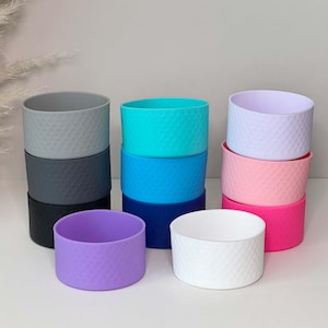 Może przedstawiać: Kolekcja kolorowych silikonowych osłon na butelki z wodą w różnych odcieniach, w tym szarym, turkusowym, niebieskim, fioletowym, różowym i białym. Każda osłona ma teksturowany wzór w romby. Osłony są ułożone w stosy, prezentując gamę dostępnych kolorów.