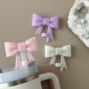 May include: Three glittery bow-shaped drink charms in pink, white, and lavender. The pink bow is attached to a light pink and gray tumbler. The charms have a sparkly finish and are set against a neutral background.