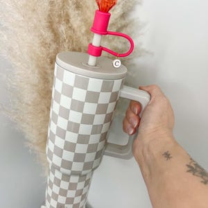May include: A large tumbler with a checkered pattern in beige and white, featuring a matching handle. It has a pink straw with a decorative orange gem at the top. The tumbler is held by a person.