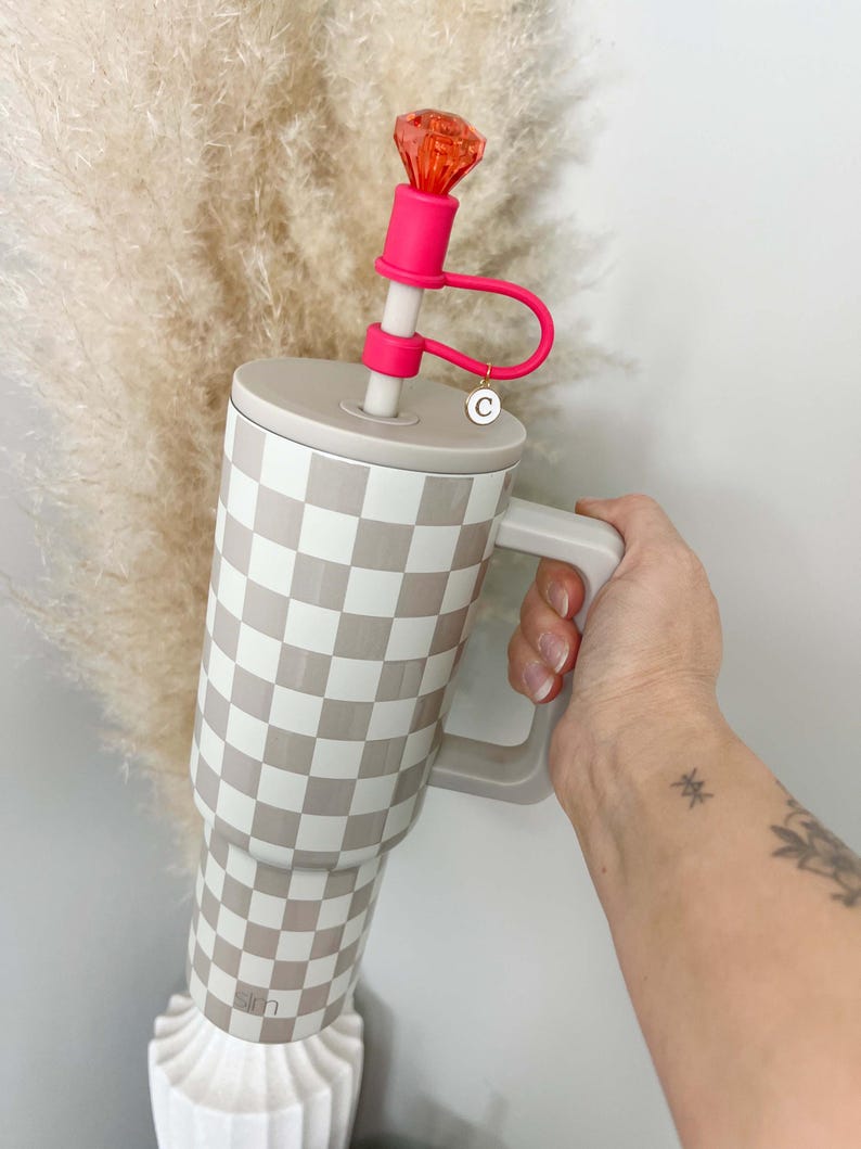 May include: A large tumbler with a checkered pattern in beige and white, featuring a matching handle. It has a pink straw with a decorative orange gem at the top. The tumbler is held by a person.